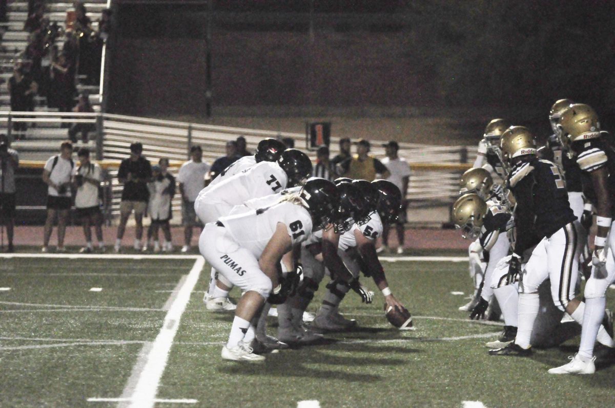 Sophomore long snapper Tanyon Erdman (#54) prepares to snap the ball to the placeholder for an extra point attempt. The offensive line is lined up and focused as well, ready to defend junior kicker James Dean.